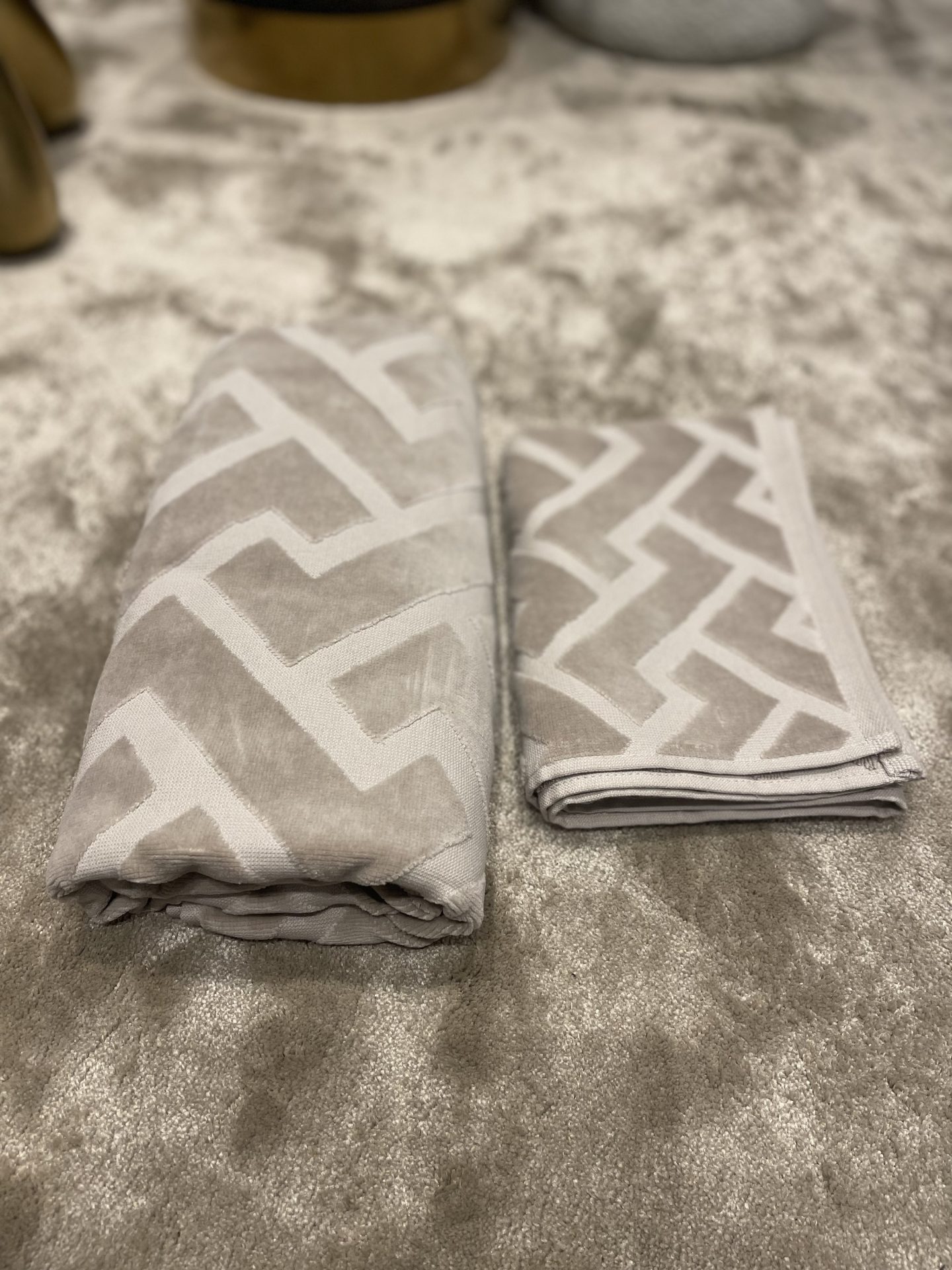 Opgevouwen badlaken en handdoek met geweven geometrisch patroon in beige, naast elkaar op een lichtgrijs vloerkleed