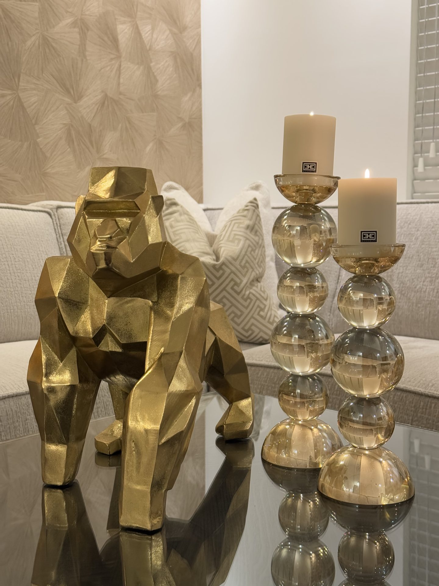Gouden geometrische gorillasculptuur op een glazen salontafel, naast twee glazen kaarshouders met stompkaarsen, voor een crèmekleurige bank