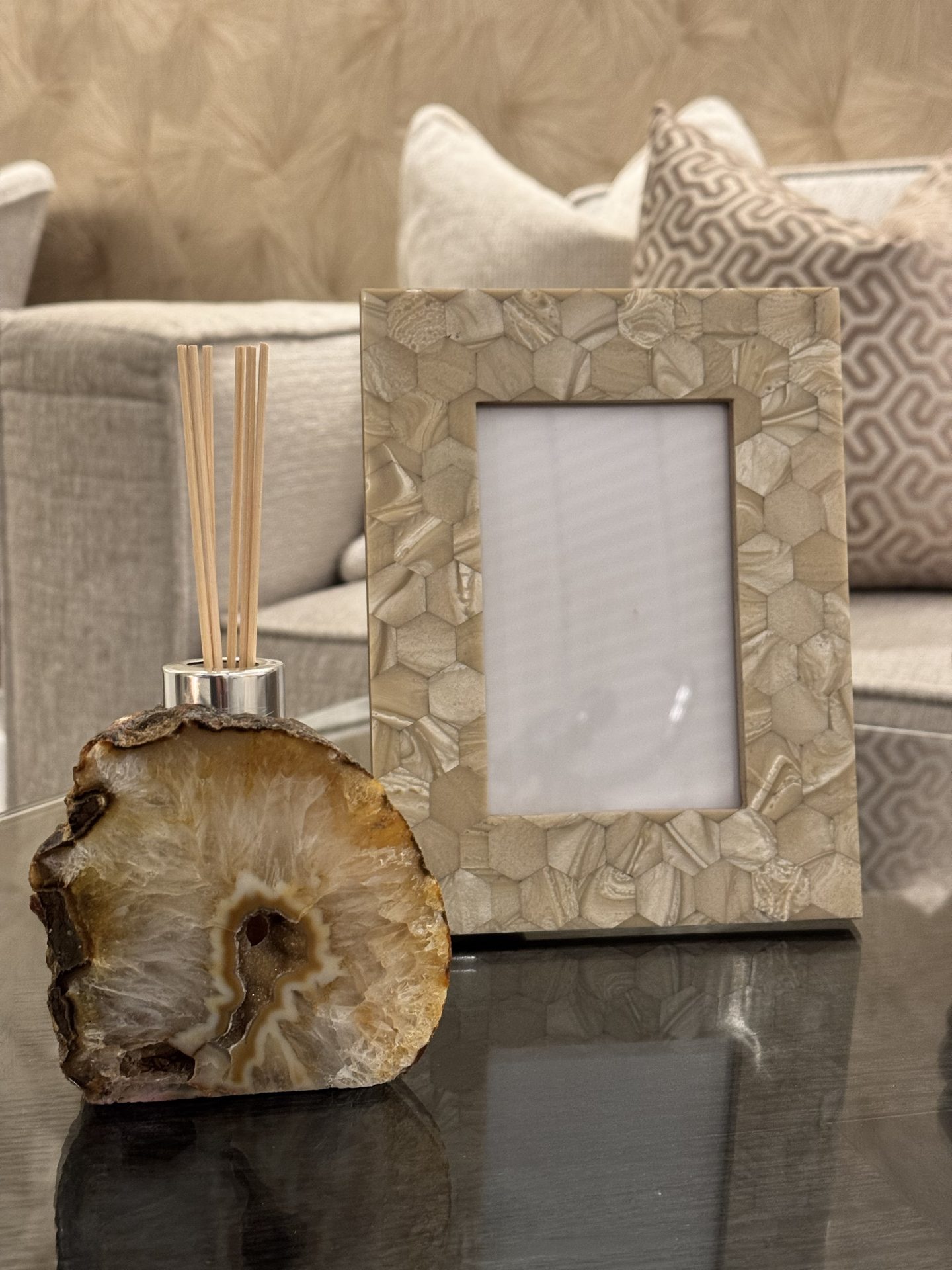 Zandkleurige fotolijst met hexagonaal schelpenpatroon naast een geurstokjeshouder van agaat op een glazen tafel