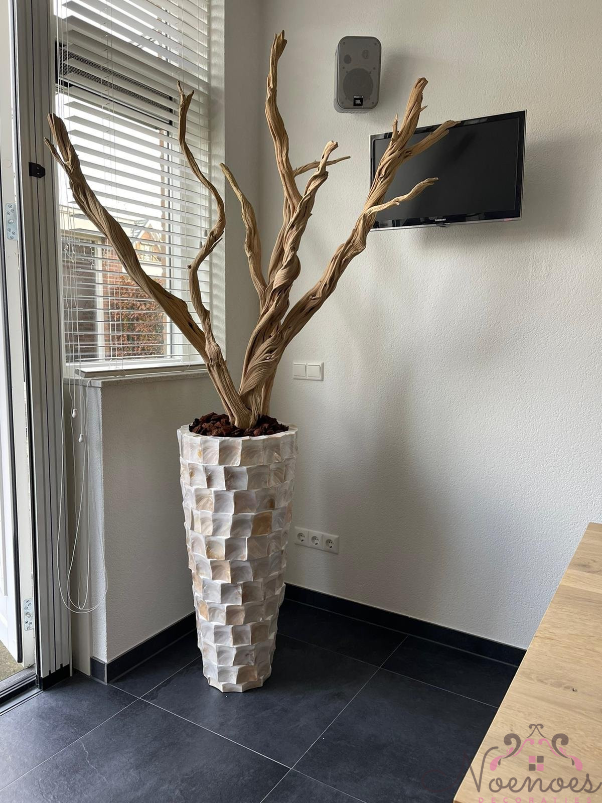 Hoge witte schelpenvaas met ghostwood takken in een hoek naast een raam met jaloezieën, tegen een lichte muur