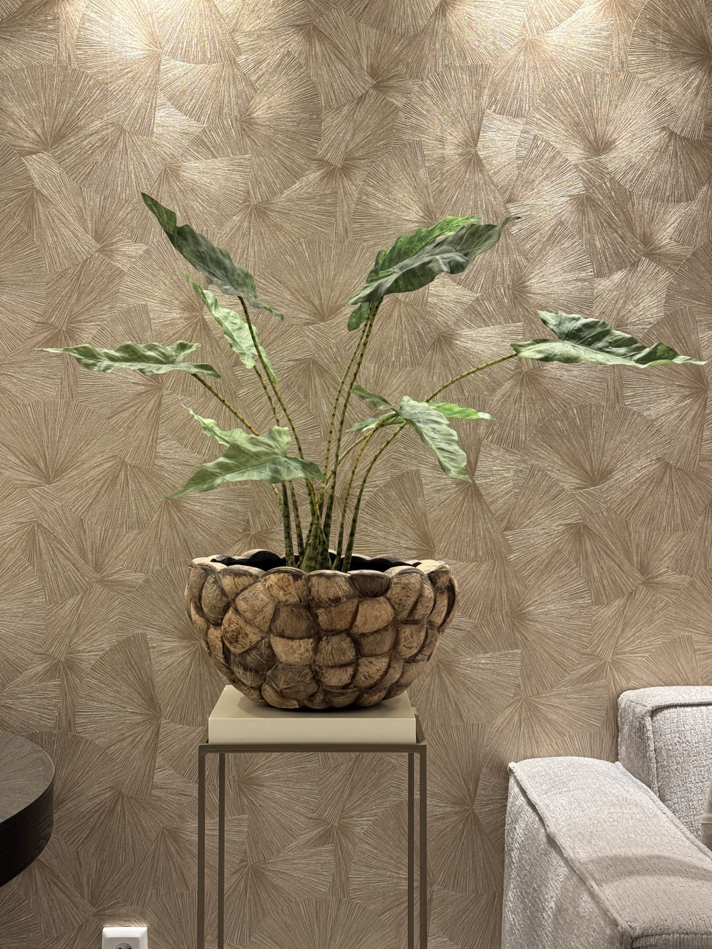 Kokosvaas met een Alocasia zebrina kunstplant op een lage sokkel tegen een wand met structuurbehang