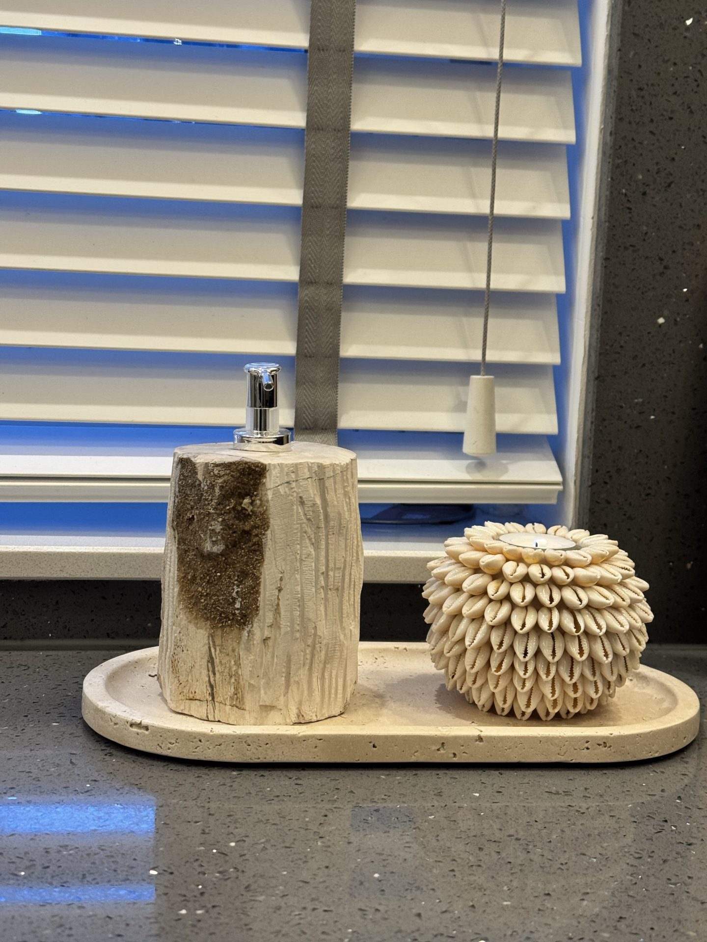 Zeeppompje van versteend hout en een met kauri-schelpen bekleed waxinelichthouder naast elkaar op een travertijnen dienblad