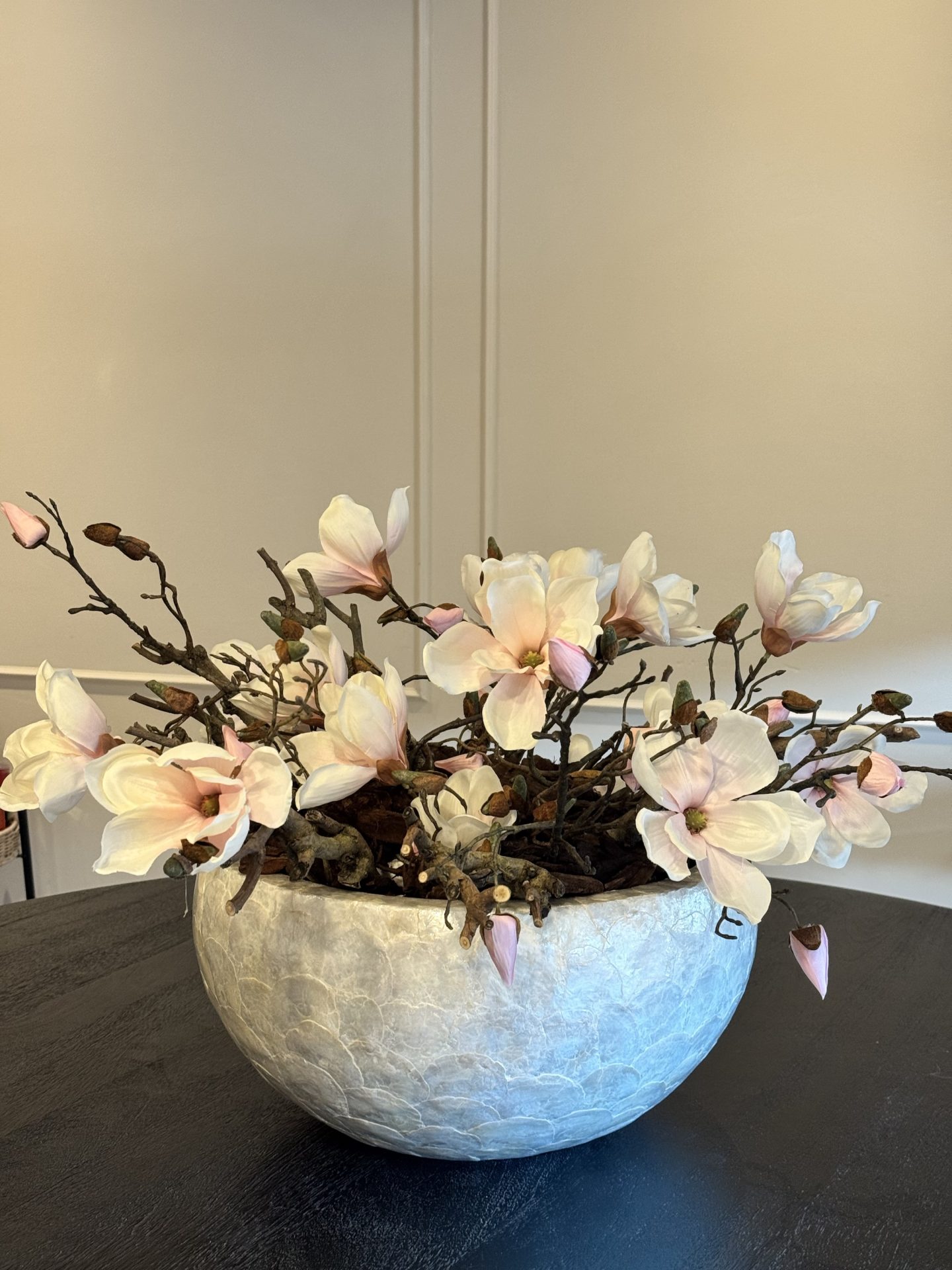 Lage schelpenvaas met zijden magnoliatakken die aan beide kanten over de rand vallen, op een donker tafelblad