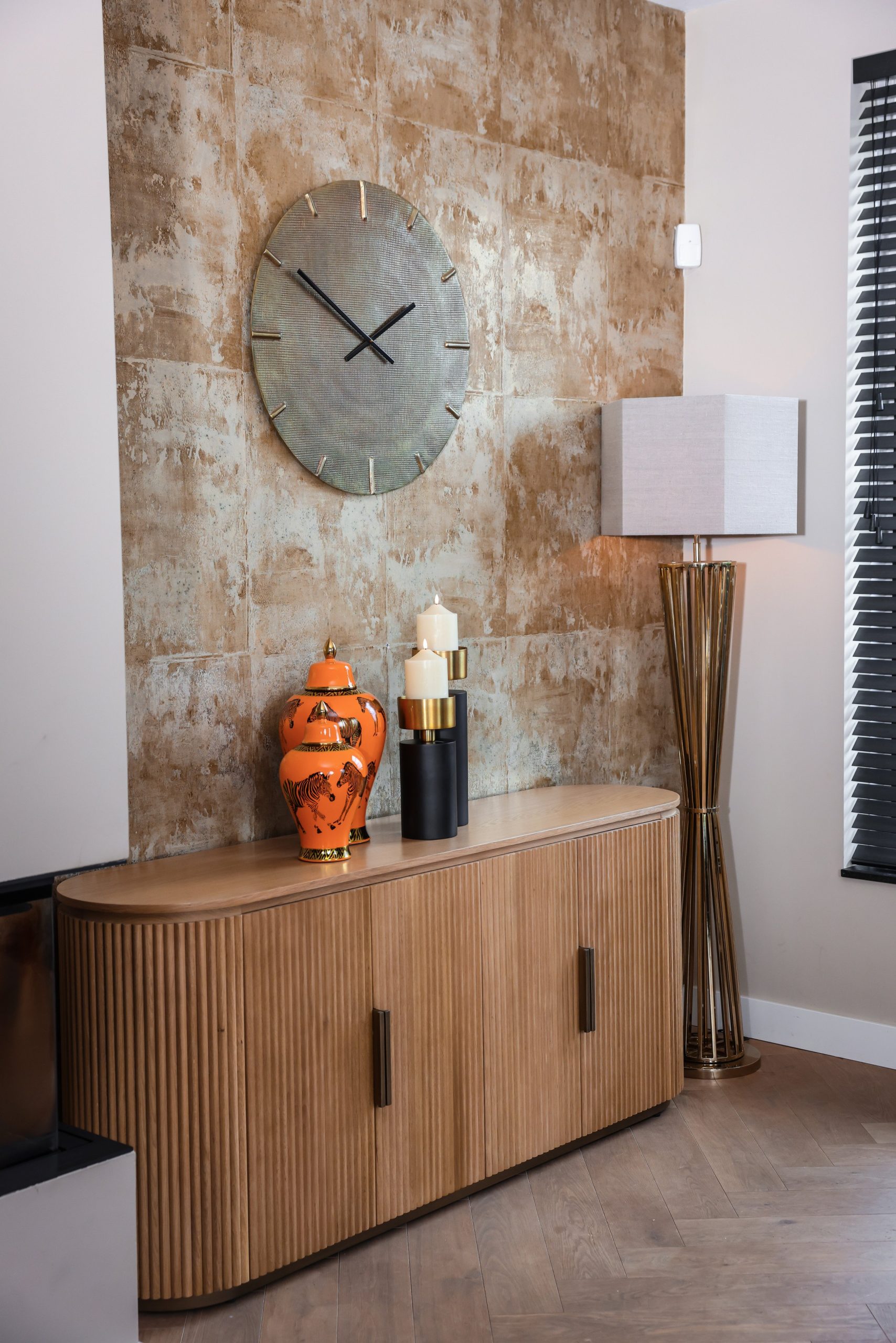 Dressoir met een oranje vaas en twee zwarte kandelaars in verschillende hoogtes tegen een verweerde wand