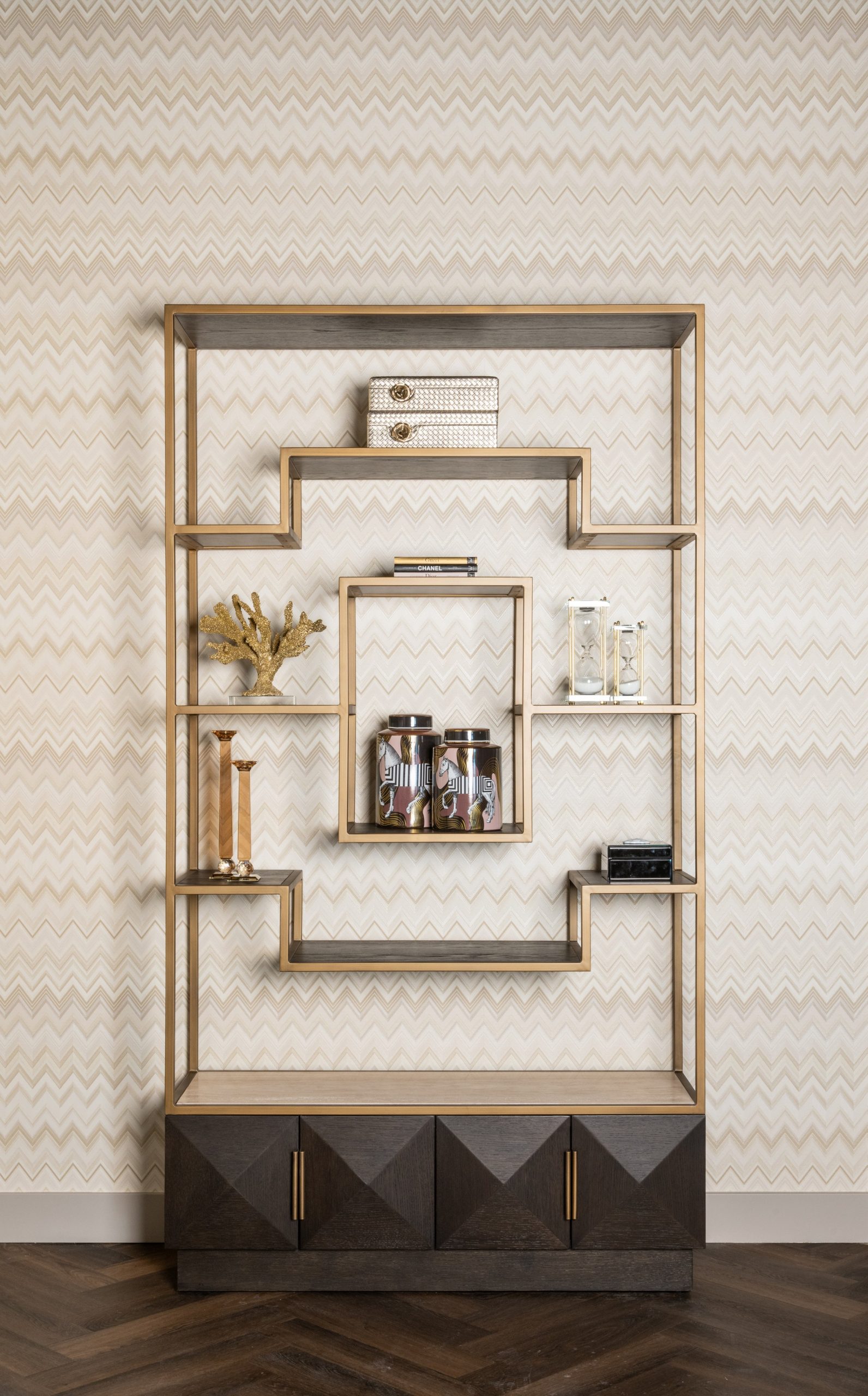 Open kast met goudkleurig frame tegen een wand met zigzagpatroon in beige, met koraal, kandelaars, potten en zandlopers op de planken