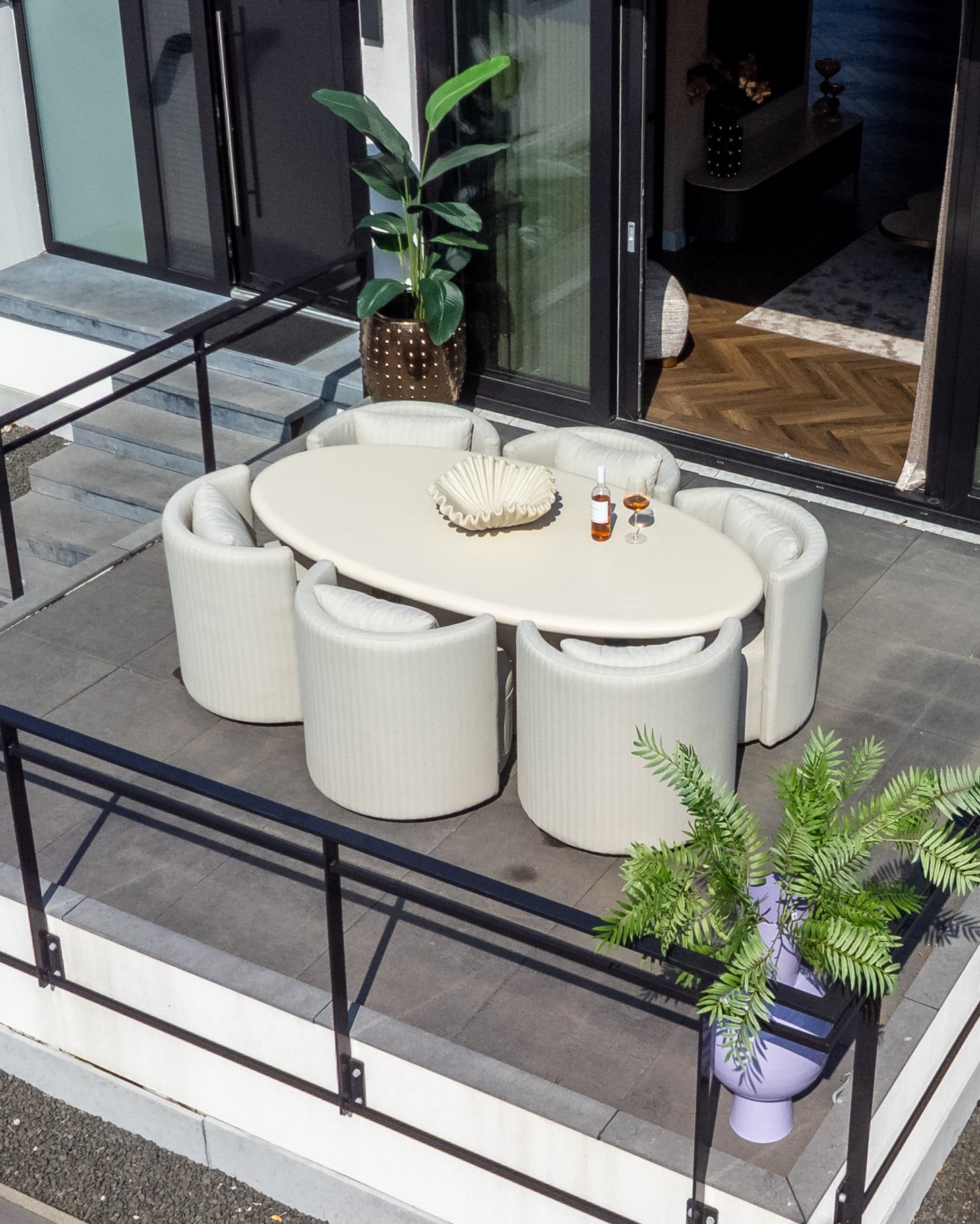 Ovale buitentafel met crèmekleurige stoelen op een terras, met een lage witte schaal, een fles wijn en twee glazen op tafel