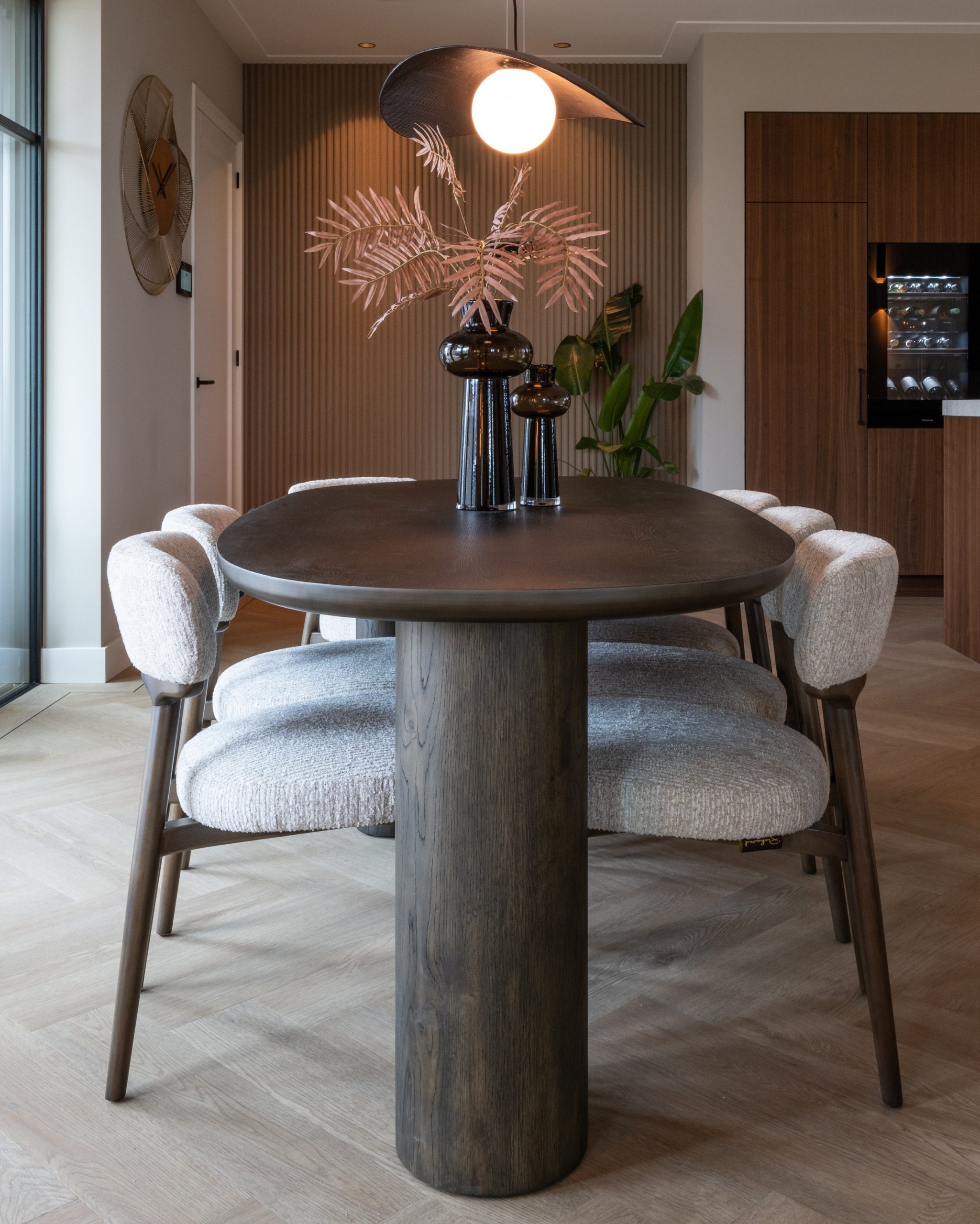 Twee rookglazen vazen in verschillende hoogtes met roze palmtakken midden op een ovale eettafel met witte bouclé stoelen