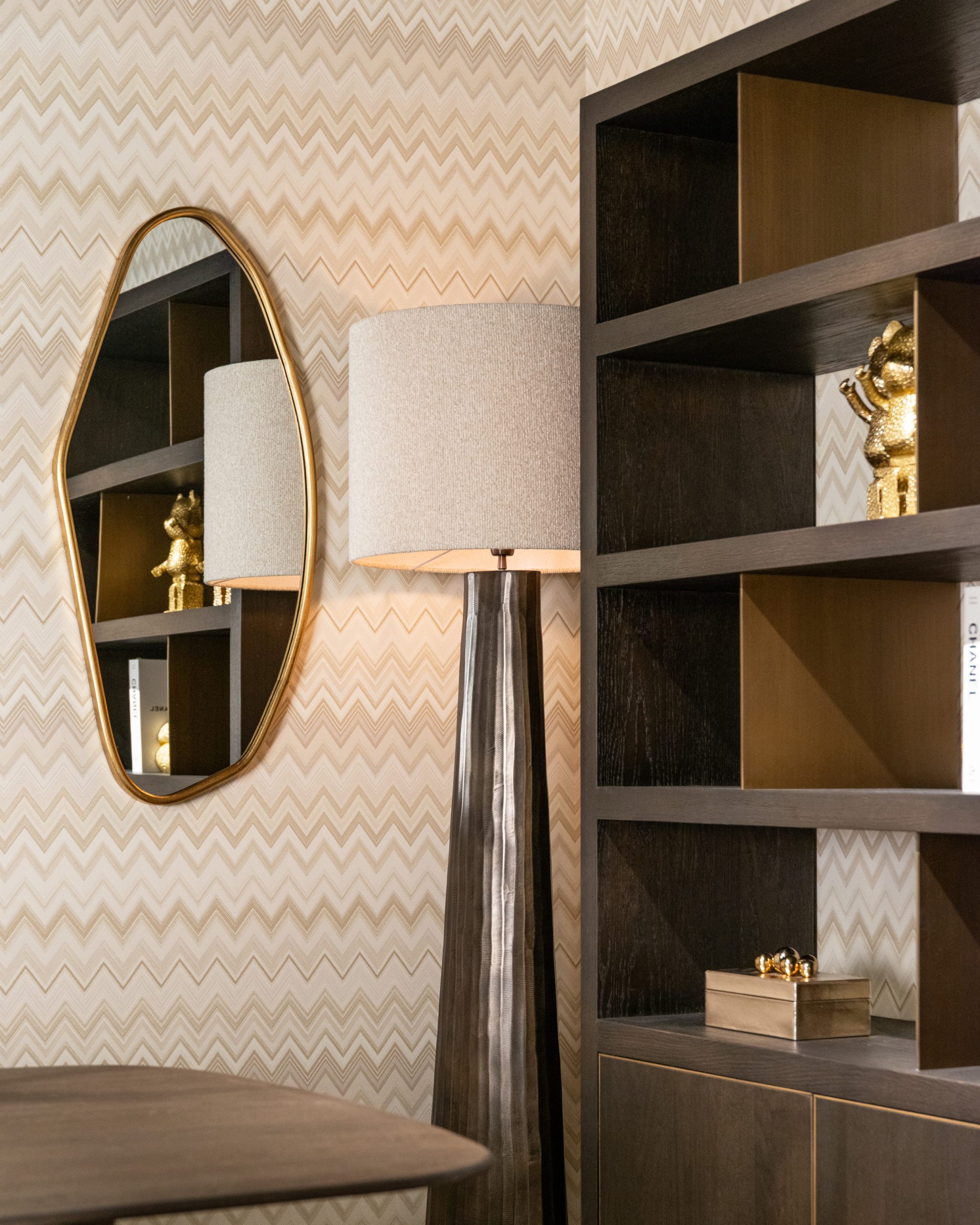 Ovale spiegel met goudkleurige lijst aan een wand met chevronpatroon, naast een aangestoken vloerlamp met linnen kap en een donkere open kast