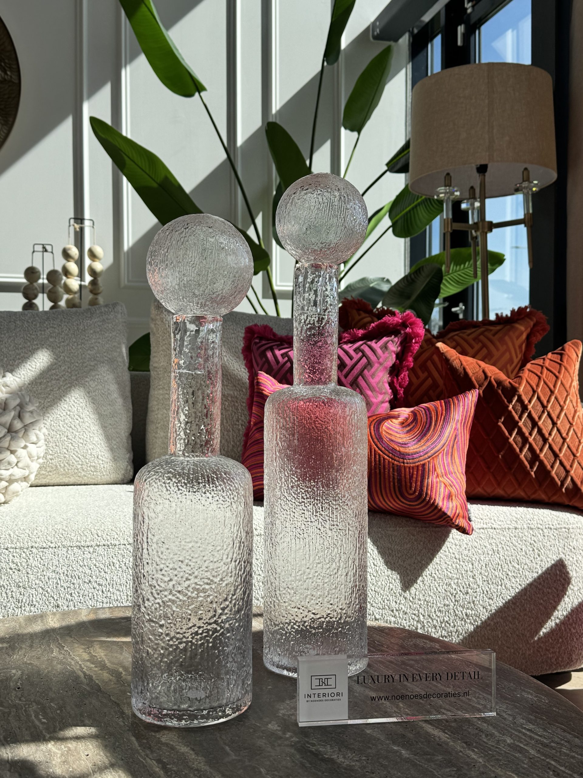 Twee hoge karaffen van transparant structuurglas met bolvormige stop in de zon, met roze en oranje sierkussens die door het glas heen zichtbaar zijn