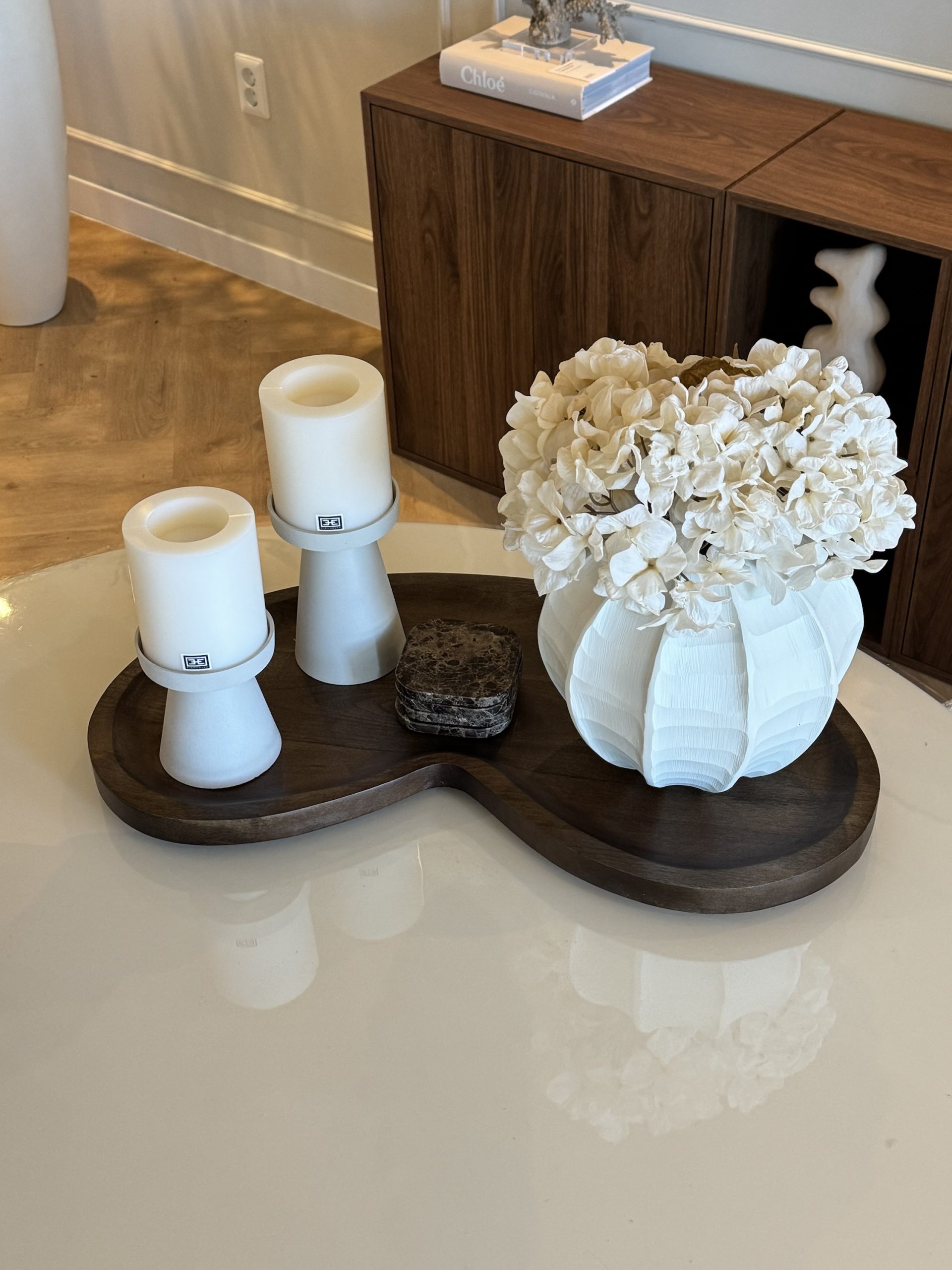 Donker houten dienblad op een lichte tafel met twee witte kaarshouders, een stapel marmeren onderzetters en een witte vaas met hortensia's