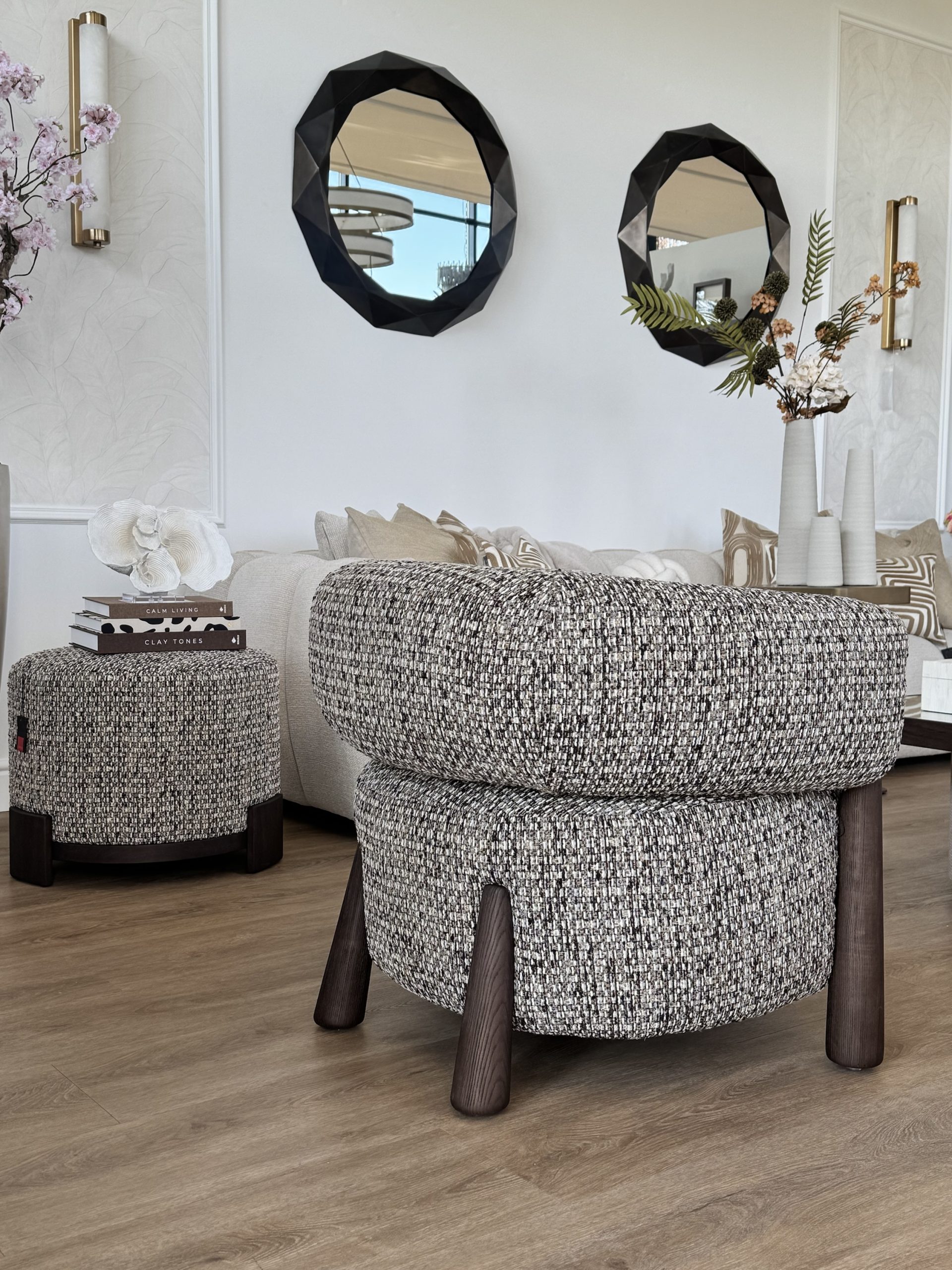 Beige poef met een stapel boeken erop, naast een bouclé fauteuil en een lichte bank met sierkussens
