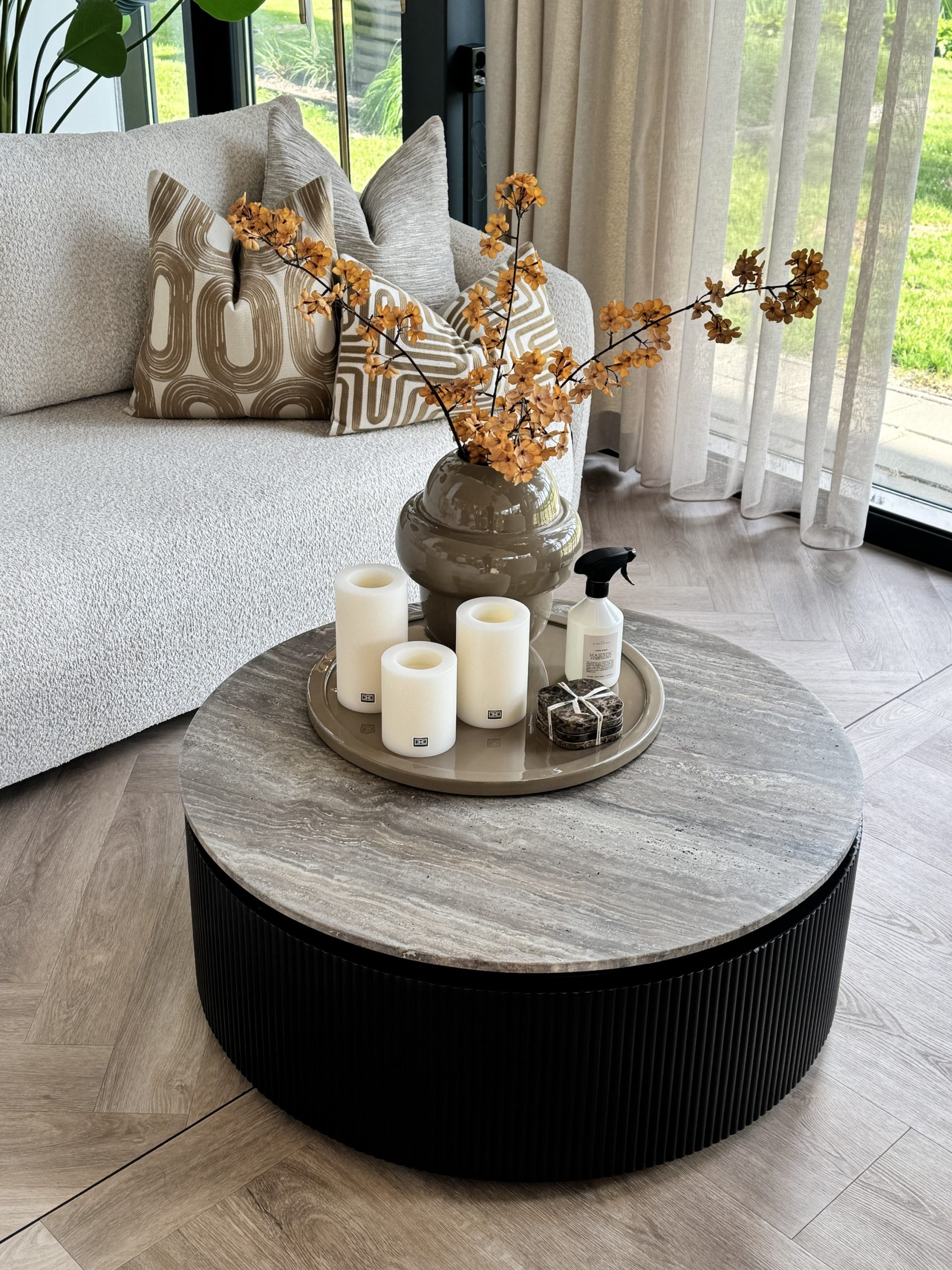 Drie witte stompkaarsen in verschillende hoogtes op een rond taupe dienblad op een salontafel, met een vaas met oranje bloesemtakken