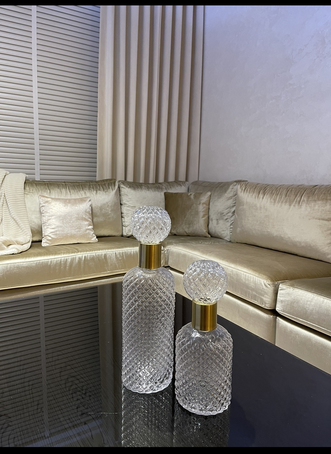 Twee glazen karaffen met reliëfpatroon en gouden stop op een tafel, met een champagnekleurige velvet hoekbank op de achtergrond